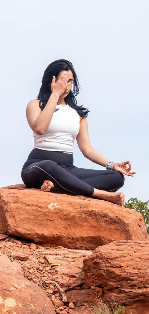 A practitioner seated cross-legged on a rock outcrop, practising alternate-nostril breathing with one hand at the nose.