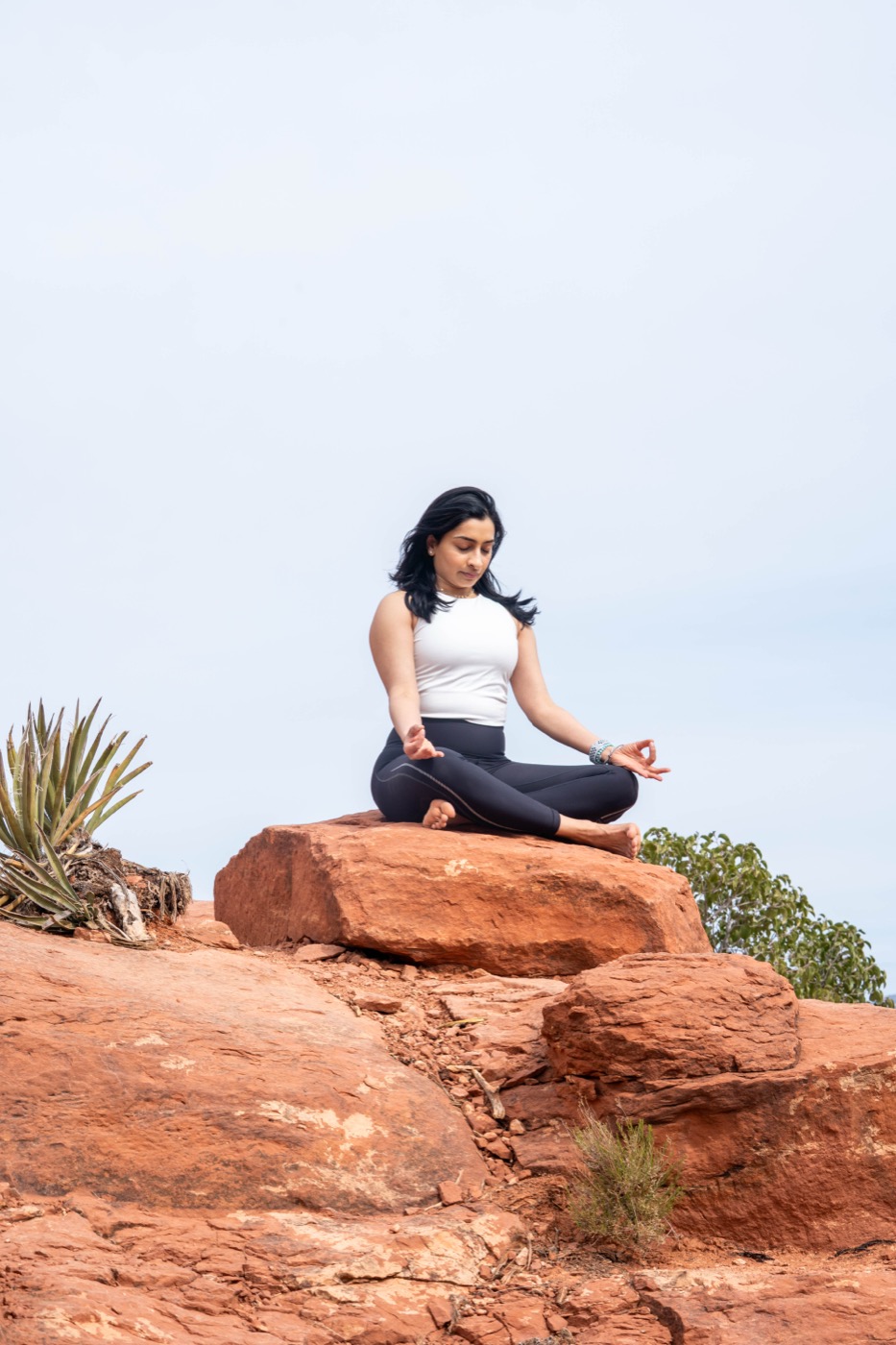 Arundhati seated cross-legged on red rock against an open sky, practising alternate-nostril breathing with one hand at her nose and the other in chin mudra.